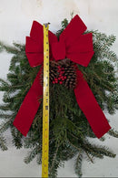 A Christmas wreath made of green pine branches with a large red fabric bow, a pinecone, and small red berries, shown against a white wall with a yellow tape measure placed vertically through the center.