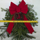 A Christmas wreath made of green pine branches with a large red bow, a pinecone, and small red berries, shown with a yellow tape measure across the front against a white background.