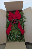 A Christmas greenery arrangement in a brown cardboard box, featuring green pine branches with a large red fabric bow, a pinecone, and small red berries.